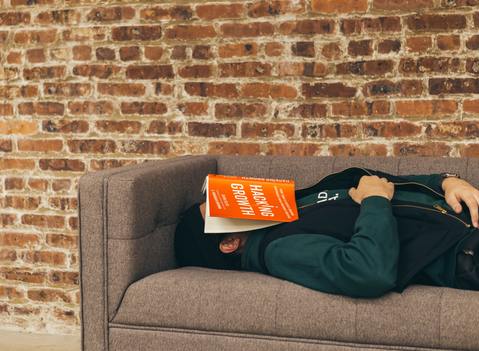 Middle-aged man in dark long sleeve shirt resting on a gray couch in front of a red brick wall after consuming cannabis for sleep with a book on his face about hacking growth boasting a bright orange cover